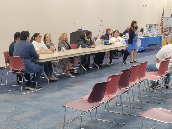 Nine people seated at a long table facing an audience inside a conference room. A woman is standing in front of the table with a mic talking to the audience.